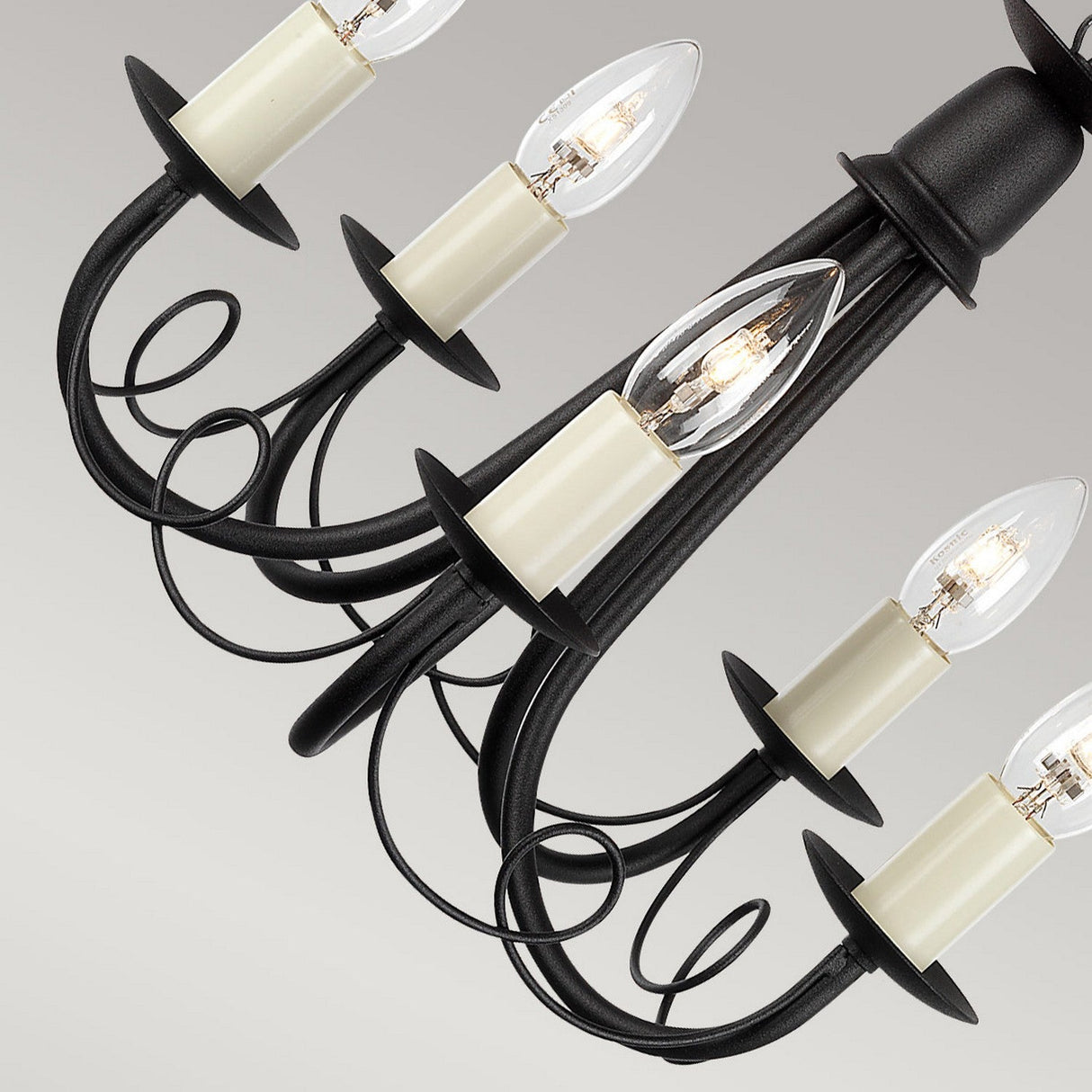 A close-up of the Minster 5 Light Chandelier in black wrought iron, showcasing its striking black finish. This sophisticated fixture includes six candle-shaped lightbulbs, each set in a cream holder. Decorative scrolls enhance its charm, all elegantly displayed against a light gray backdrop for the perfect blend of elegance and style.