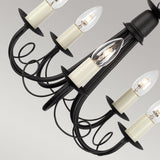 A close-up of the Minster 5 Light Chandelier in black wrought iron, showcasing its striking black finish. This sophisticated fixture includes six candle-shaped lightbulbs, each set in a cream holder. Decorative scrolls enhance its charm, all elegantly displayed against a light gray backdrop for the perfect blend of elegance and style.