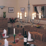 A dining room exudes warmth with a wooden table set for a meal. Plates, glasses, and a bottle of red wine complement the flickering candles. Above, the Minster 6 Light Chandelier in black and gold patina casts an inviting glow, while cabinets and framed art complete the elegant ambiance.