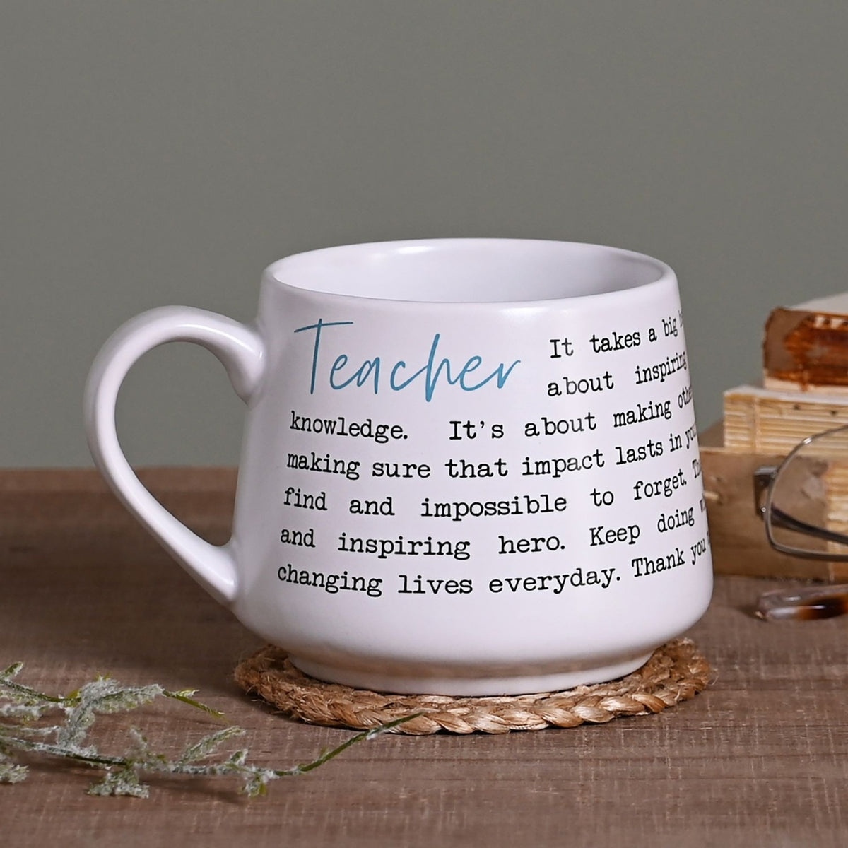 White stoneware mug with C-shaped handle, featuring "Teacher" in blue script and a black typewriter-style thank-you verse, shown on a wooden table with a woven coaster.