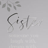Close-up of the grey hinged photo frame's right panel, showing "Sister" in silver script above a white verse beginning "someone you laugh with, dream with," with a subtle leaf motif in the corner.