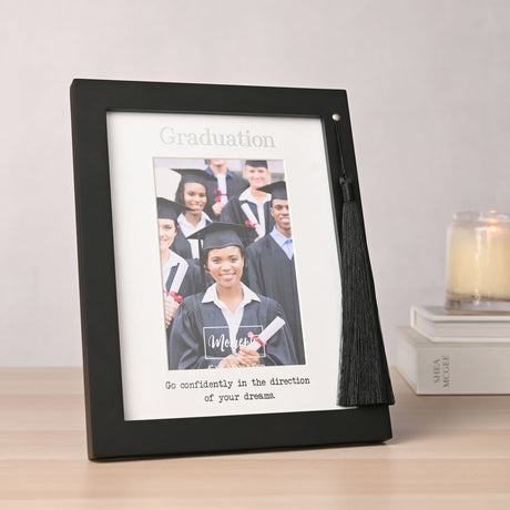 Black wooden 6x4in Graduation photo frame with white mount and black tassel, shown on a wooden table beside books and a candle.