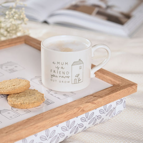White stoneware mug with frothy drink, printed "a MUM is a FRIEND you never OUT GROW" and a house and flower design, shown on a wooden tray with cookies.