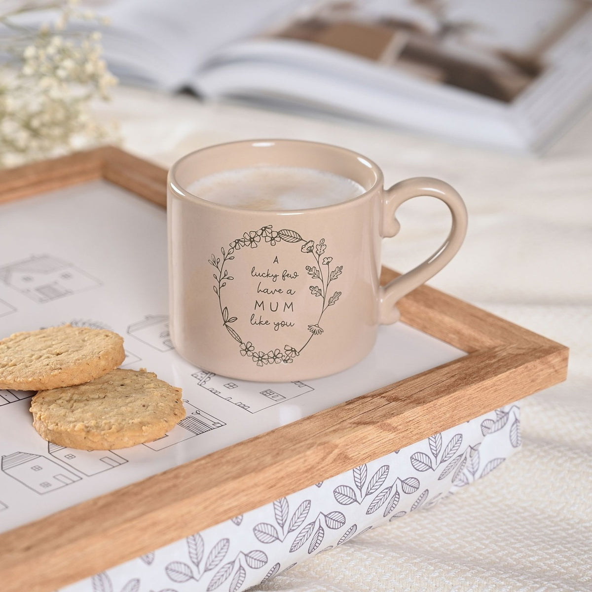 Brown stoneware mug with curled handle and floral wreath design reading "A lucky few have a MUM like you", filled with coffee on a wooden tray beside biscuits.
