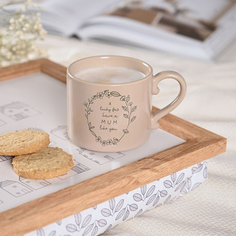 Brown stoneware mug with curled handle and floral wreath design reading "A lucky few have a MUM like you", filled with coffee on a wooden tray beside biscuits.
