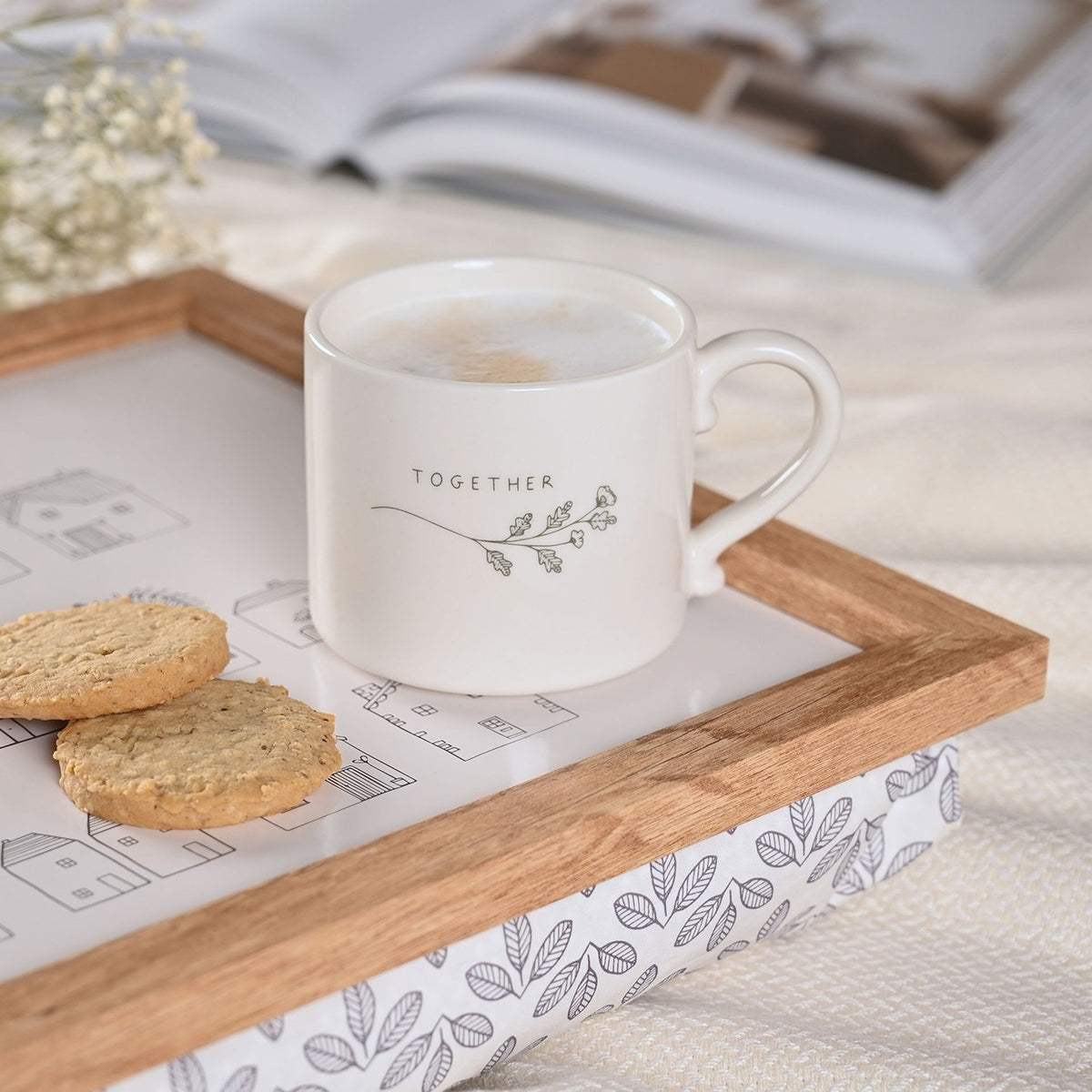 White stoneware mug with curled handle, printed "TOGETHER" text and a grey floral sprig, filled with coffee on a wooden tray beside biscuits.