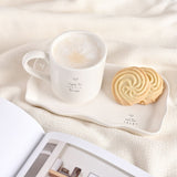 Off-white stoneware mug with heart print and "enjoy the LITTLE things" text, paired with a wavy-edged rectangular plate holding swirl biscuits, on a cream fabric surface.