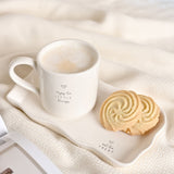Off-white stoneware mug filled with frothy coffee, printed with a heart and "enjoy the LITTLE things," set on a matching wavy-edged rectangular plate reading "eat the TREAT" beside two swirl biscuits.