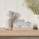 Grey wooden mantel plaque with three line-drawn houses on a natural wood base reading "Friends Are The Family We Choose," shown on a shelf with mugs and a vase of flowers.