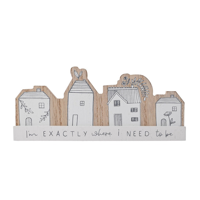 Wooden mantel plaque with four white house silhouettes in grey linework and floral details, on a base reading "I'm EXACTLY where i NEED to be", set against a plain white background.
