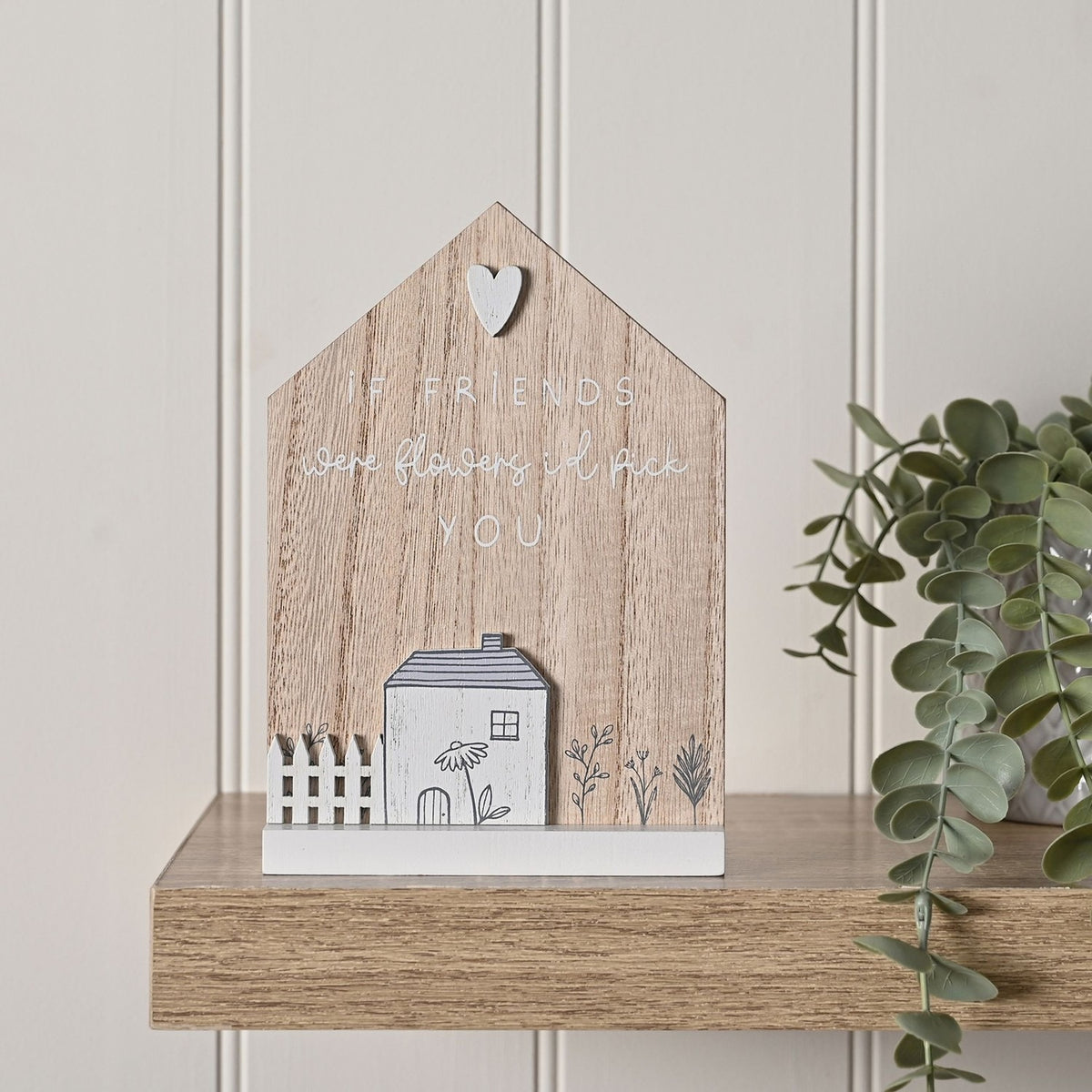 Wooden house-shaped plaque with pointed roof and white heart, reading "If friends were flowers I'd pick you" above a white cottage, fence, and florals, displayed on a wood shelf near a plant.
