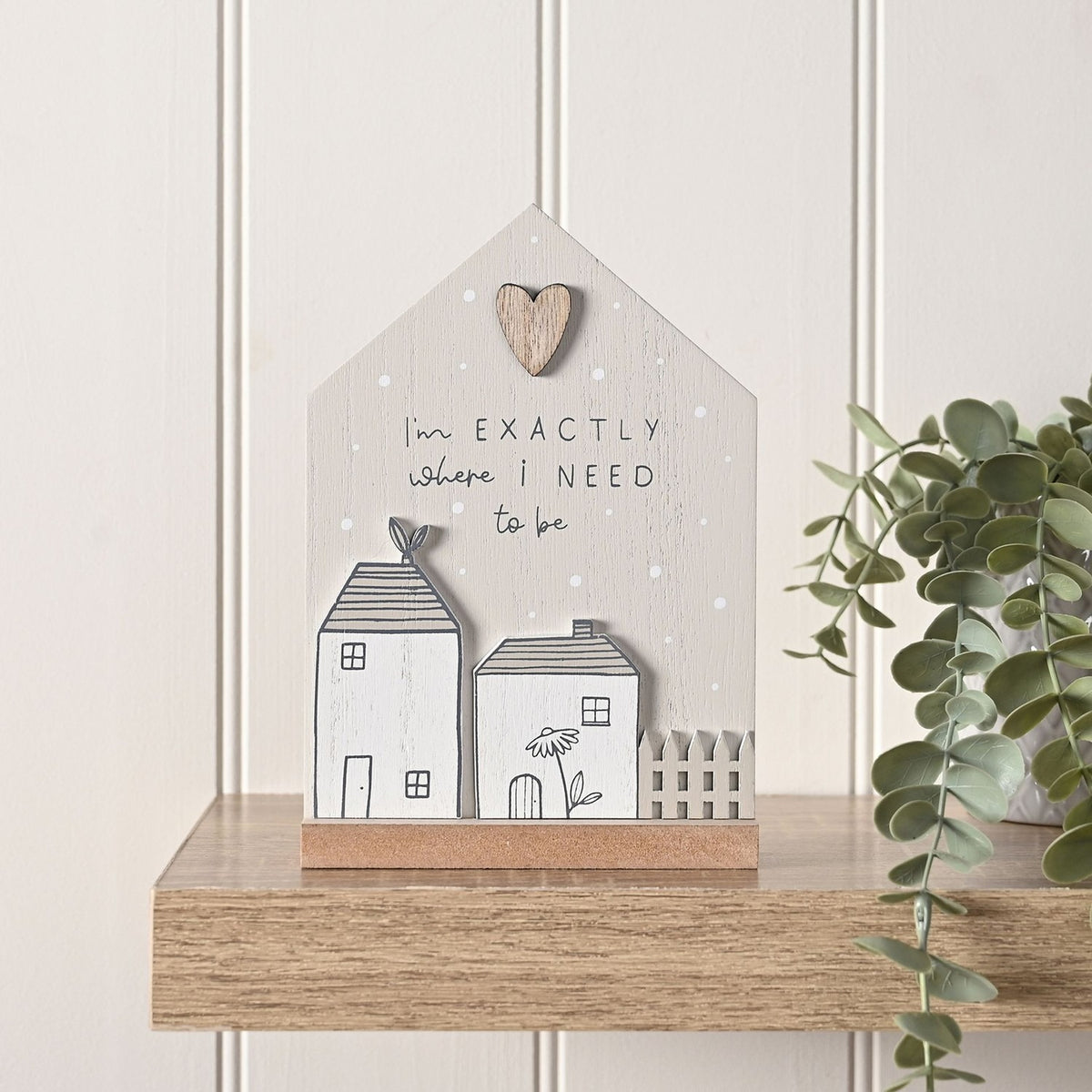Grey wooden house-shaped plaque reading "I'm EXACTLY where i NEED to be" with raised white cottage cutouts and a small wooden heart, displayed on a wooden shelf.