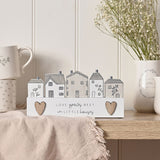 Love Grows Little Houses Wooden Plaque with Hearts in White, featuring five hand-drawn houses and wooden hearts, displayed on a shelf beside mugs and a vase of flowers.
