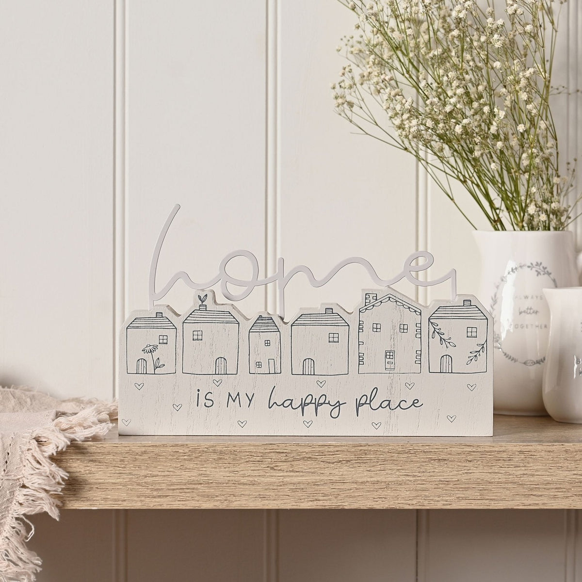 Wooden mantel plaque in grey with six line-drawn houses and "home IS MY happy place" script, shown on a wooden shelf beside a white vase of flowers.