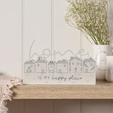 Wooden mantel plaque in grey with six line-drawn houses and "home IS MY happy place" script, shown on a wooden shelf beside a white vase of flowers.
