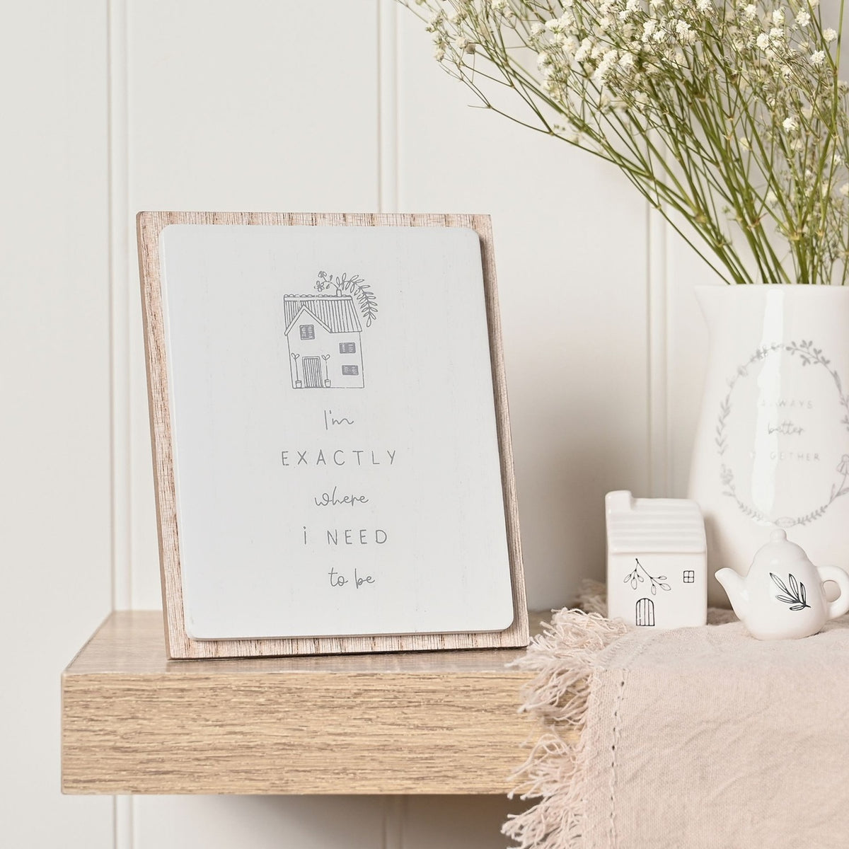 Layered wooden wall plaque with white glazed panel, grey house illustration, and I'm Exactly Where I Need to Be text, shown on a shelf beside a white jug of flowers.