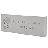 White wooden mantel plaque with a grey house line drawing and "I'm EXACTLY where i NEED to be" text, shown at an angle on a plain background.