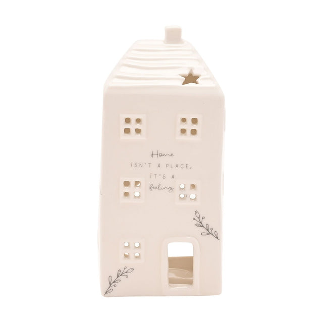White ceramic cut-out house tea light candle holder with ridged roof, chimney, star cut-out, pierced windows, and "Home isn't a place, it's a feeling" text, on a plain background.