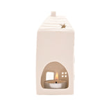 White ceramic house-shaped tea light candle holder with ridged roof, chimney, star cut-out, and arched front opening revealing a lit tea light, shown on a plain white background.