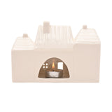 White ceramic tea light candle holder shaped like three terraced houses with ribbed roofs, chimneys, and cut-out windows, lit tealight glowing inside on a plain background.