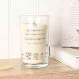 Lit tall scented candle in a clear glass jar with cream wax, printed with a heart, "friends" sentiment text, and a row of line-drawn houses, set on a wooden surface beside matches and a book.