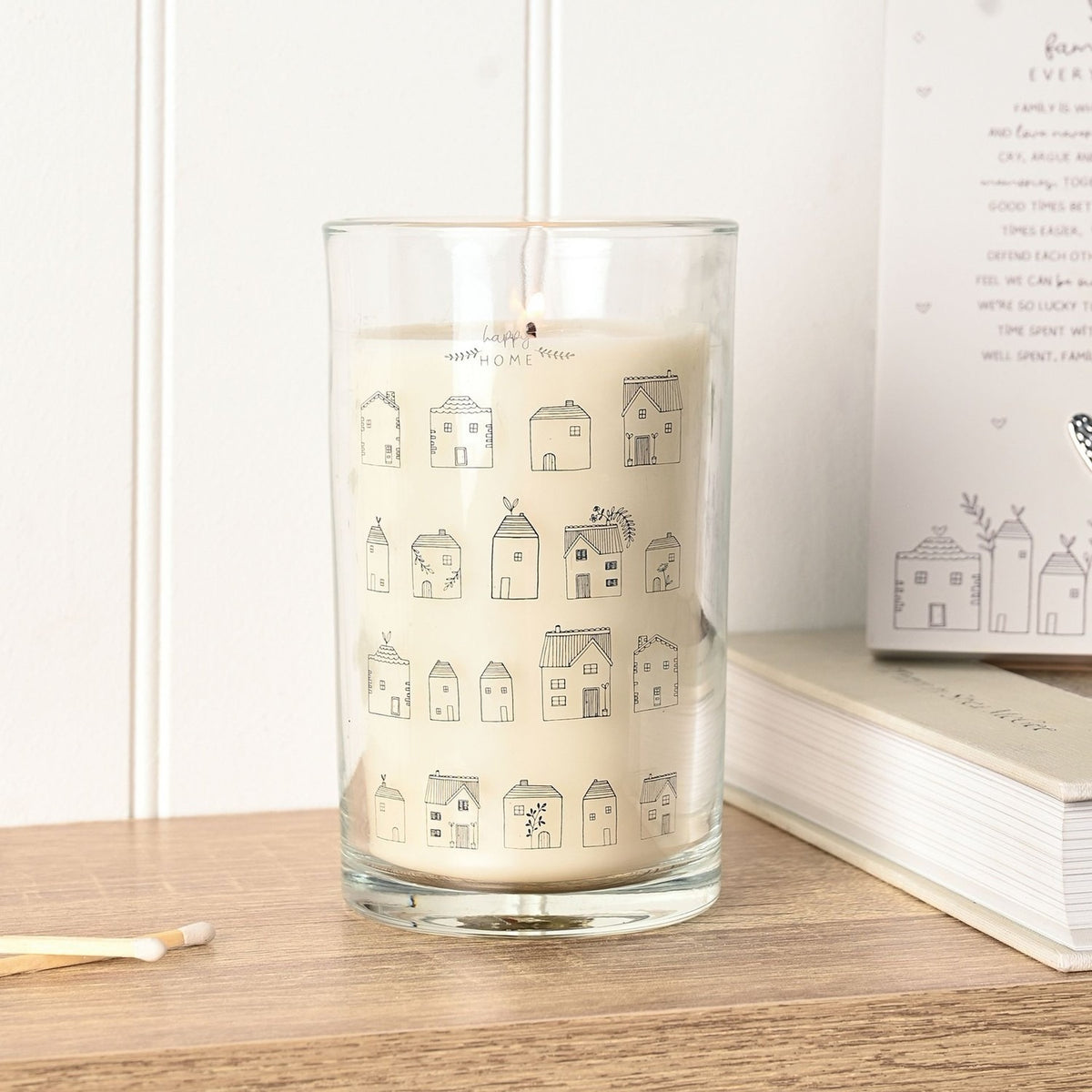 Tall scented candle in a clear glass jar with cream wax, featuring hand-drawn grey house illustrations and happy HOME lettering, lit on a wooden surface beside matches and a book.