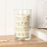 Tall scented candle in a clear glass jar with cream wax, featuring hand-drawn grey house illustrations and happy HOME lettering, lit on a wooden surface beside matches and a book.