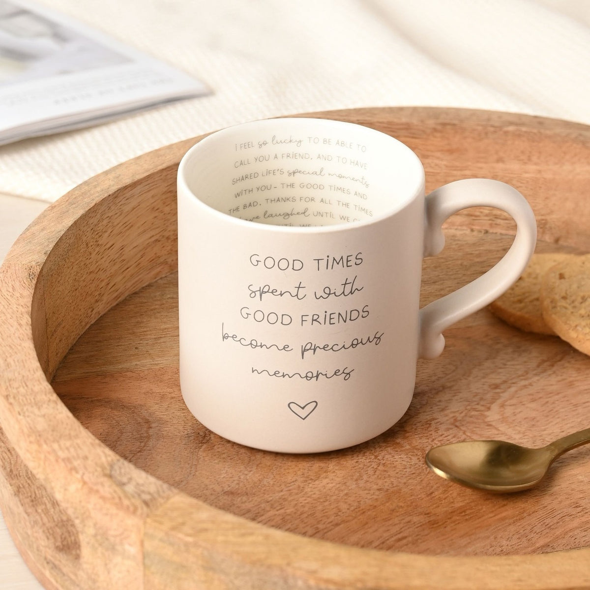 Cream ceramic mug with grey printed text "Good times spent with good friends become precious memories" and a heart outline, plus hidden lettering inside the rim, shown in a wooden tray with a spoon.
