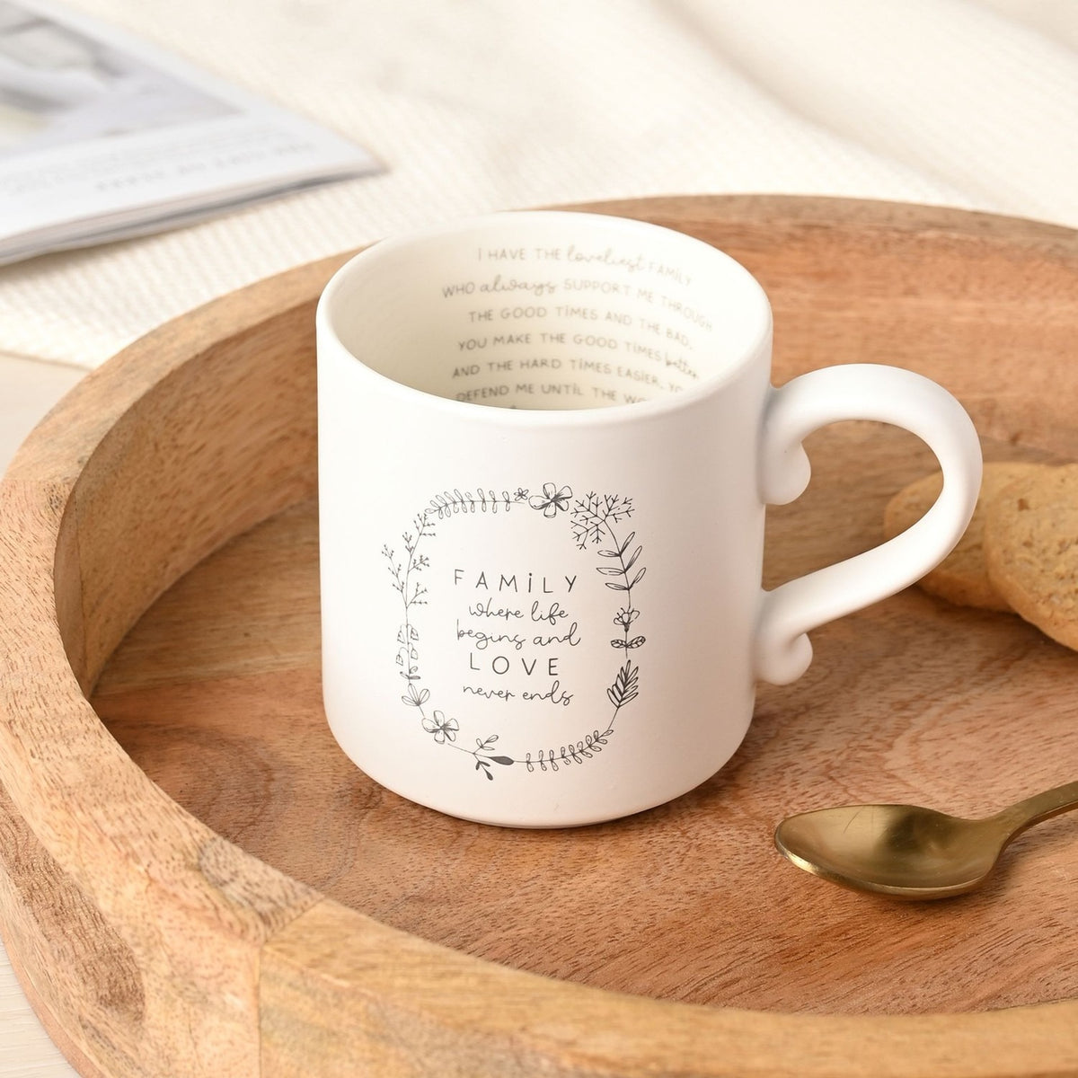 Cream ceramic mug with a grey floral wreath design and "FAMILY where life begins and LOVE never ends" text, plus an inner verse, shown in a wooden tray.