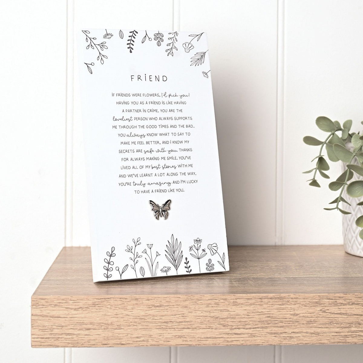 Off white rectangular standing plaque with "Friend" verse in grey text, botanical line illustrations, and a small butterfly accent, shown on a wooden shelf beside a potted plant.