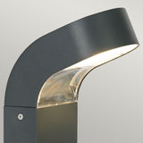Introducing the Molde LED Bollard - Graphite, a stylish outdoor lamp made from die-cast aluminium featuring a sleek, curved design. Its angled top section emits a soft white light that creates an elegant look, perfect for illuminating outdoor pathways against neutral backdrops.