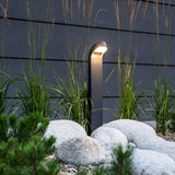 The Molde LED Bollard in Graphite casts a contemporary glow over a garden adorned with tall grasses and smooth white stones. Crafted from die-cast aluminum, it stands elegantly against a dark paneled wall, enhancing the serene ambiance.