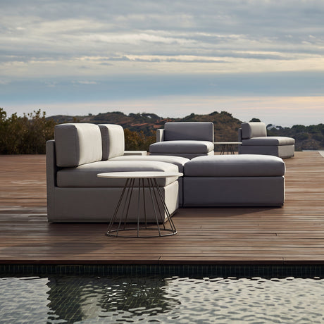 Mood XL upholstered garden corner sofa module in light grey fabric, shown with matching ottoman and round side table on a wooden poolside deck.