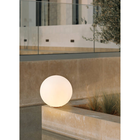 Mosvine Bollard Light in white, a spherical plastic globe on a low round base, glowing on a stone ledge beside an outdoor planted area at dusk.