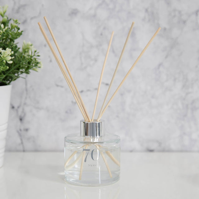 Round clear glass reed diffuser with silver "70 Seventy" foiled lettering and silver collar, reeds fanned out, set on a surface against a marble background with a plant nearby.