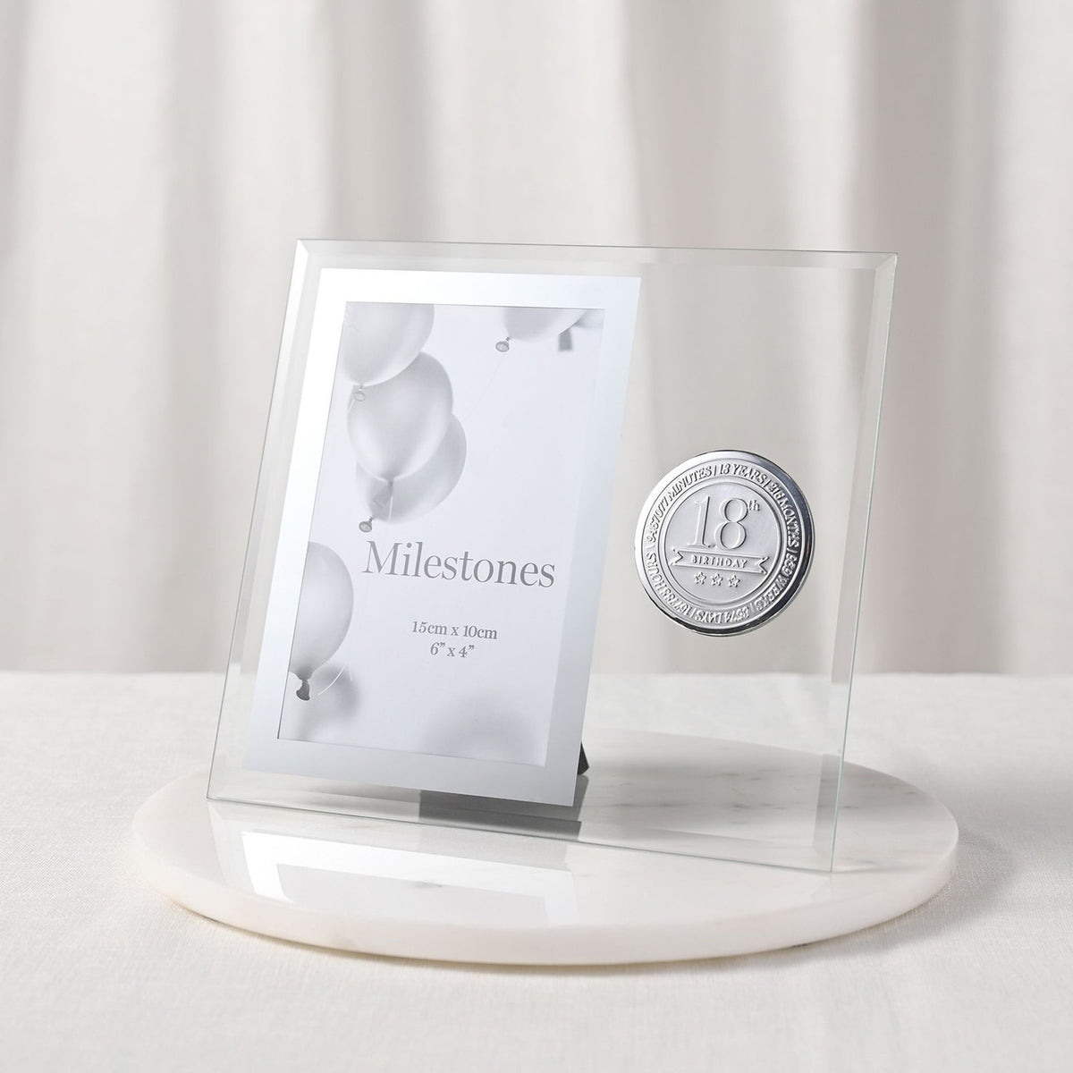 Clear glass photo frame for a 6 x 4in picture, with bevelled edges and a round silver 18th Birthday badge, standing on a white marble base.