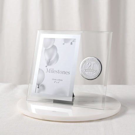 Clear glass 6x4in photo frame with round silver "21st Birthday" metal badge beside the picture opening, set on a white marble base against a light curtain background.