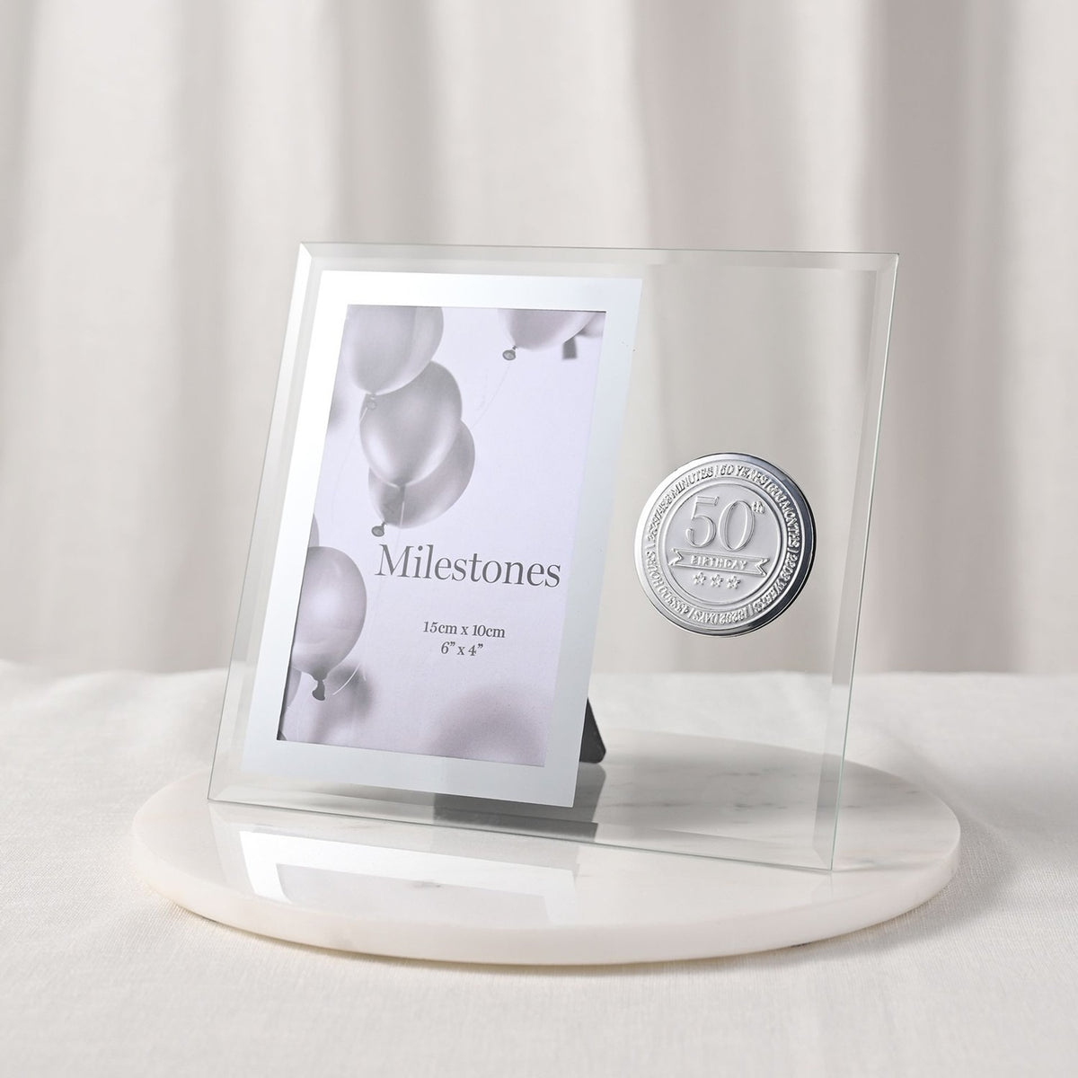 Glass photo frame with silver 50th birthday metal badge beside a 6 x 4in portrait opening, clear glass and silver-grey border, displayed on a marble base against a light background.