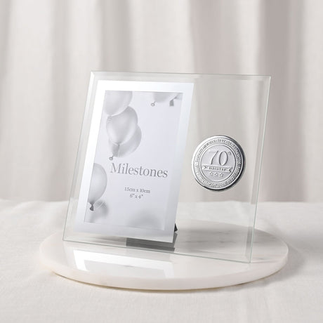 70th Birthday Metal Badge Glass Frame, 6 x 4in clear glass photo frame with silver border and round silver badge, set on a marble base against a light background.
