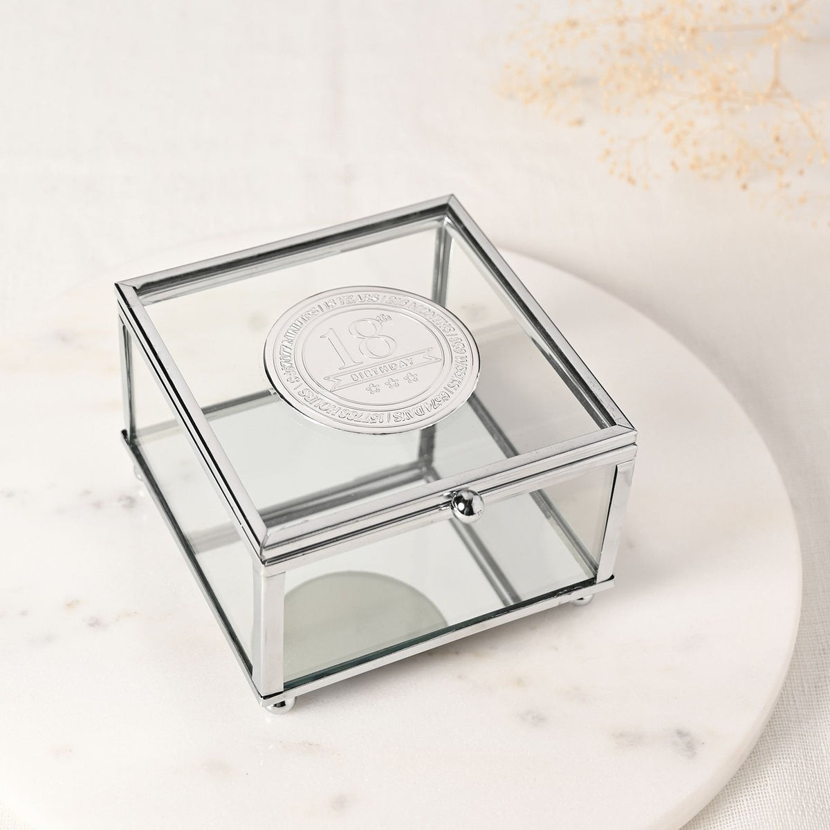Square glass trinket box with slim silver metal frame and ball feet, featuring a round silver badge on the lid engraved "18th Birthday" with stars, shown on a marble surface.