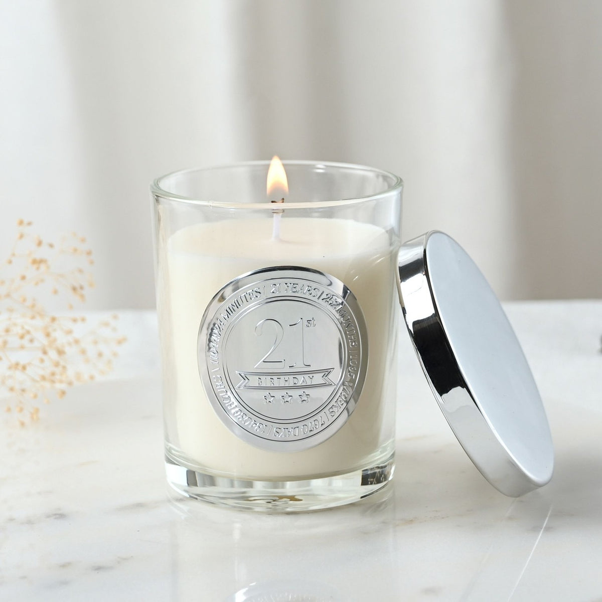 Lit 21st Birthday sandalwood scented candle in a glass jar with cream wax, silver medallion, and matching lid, set on a marble surface.