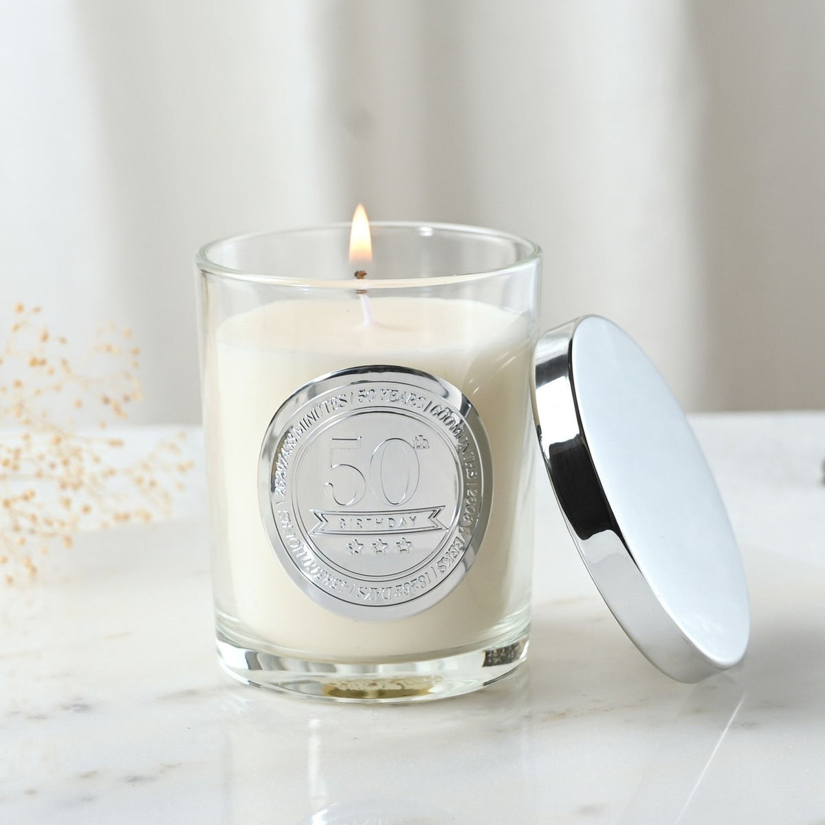 Lit 50th Birthday sandalwood scented candle in a clear glass jar with round silver medallion, shown with its polished silver lid beside it on a marble surface.