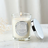 Lit 50th Birthday sandalwood scented candle in a clear glass jar with round silver medallion, shown with its polished silver lid beside it on a marble surface.
