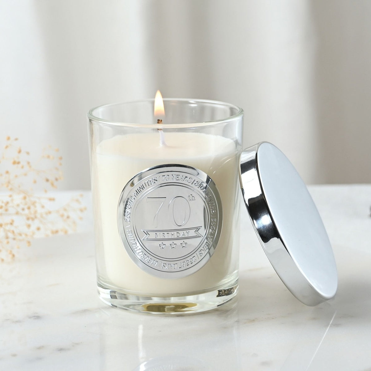 Lit 70th Birthday sandalwood scented candle in a clear glass jar with round silver medallion, shown with its silver lid beside it on a marble surface.