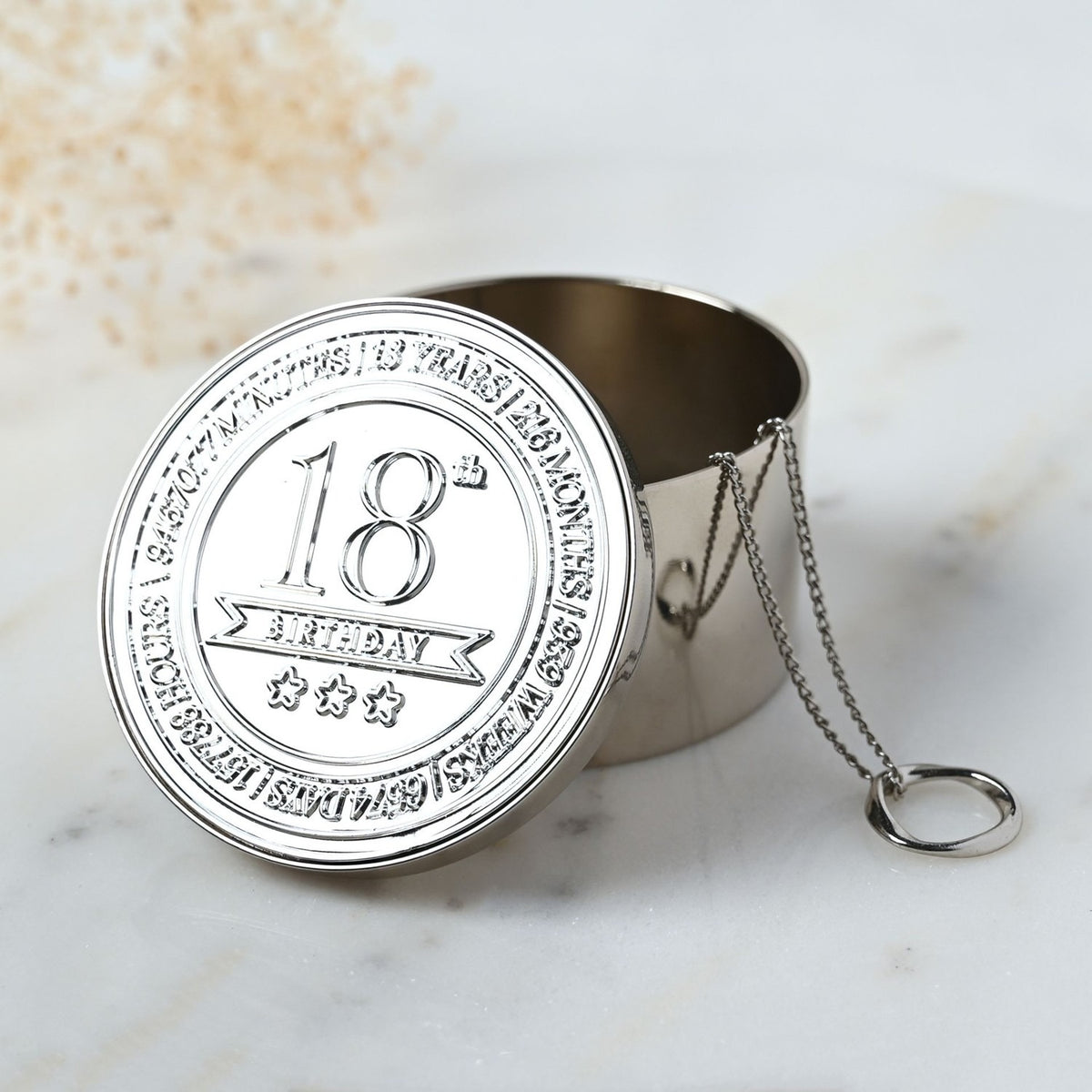 Round silver metal trinket box, lid open, embossed "18th Birthday" with stars and banner design, delicate chain hinge, shown on a marble surface.