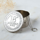 Round silver metal trinket box, lid open, embossed "18th Birthday" with stars and banner design, delicate chain hinge, shown on a marble surface.