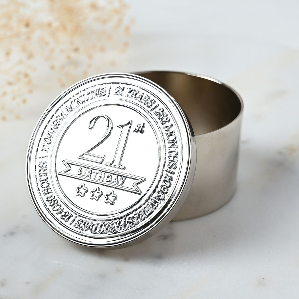 Round silver metal trinket box with open lid embossed "21st Birthday" and three stars, bordered by a time-count inscription, shown on a marble surface.