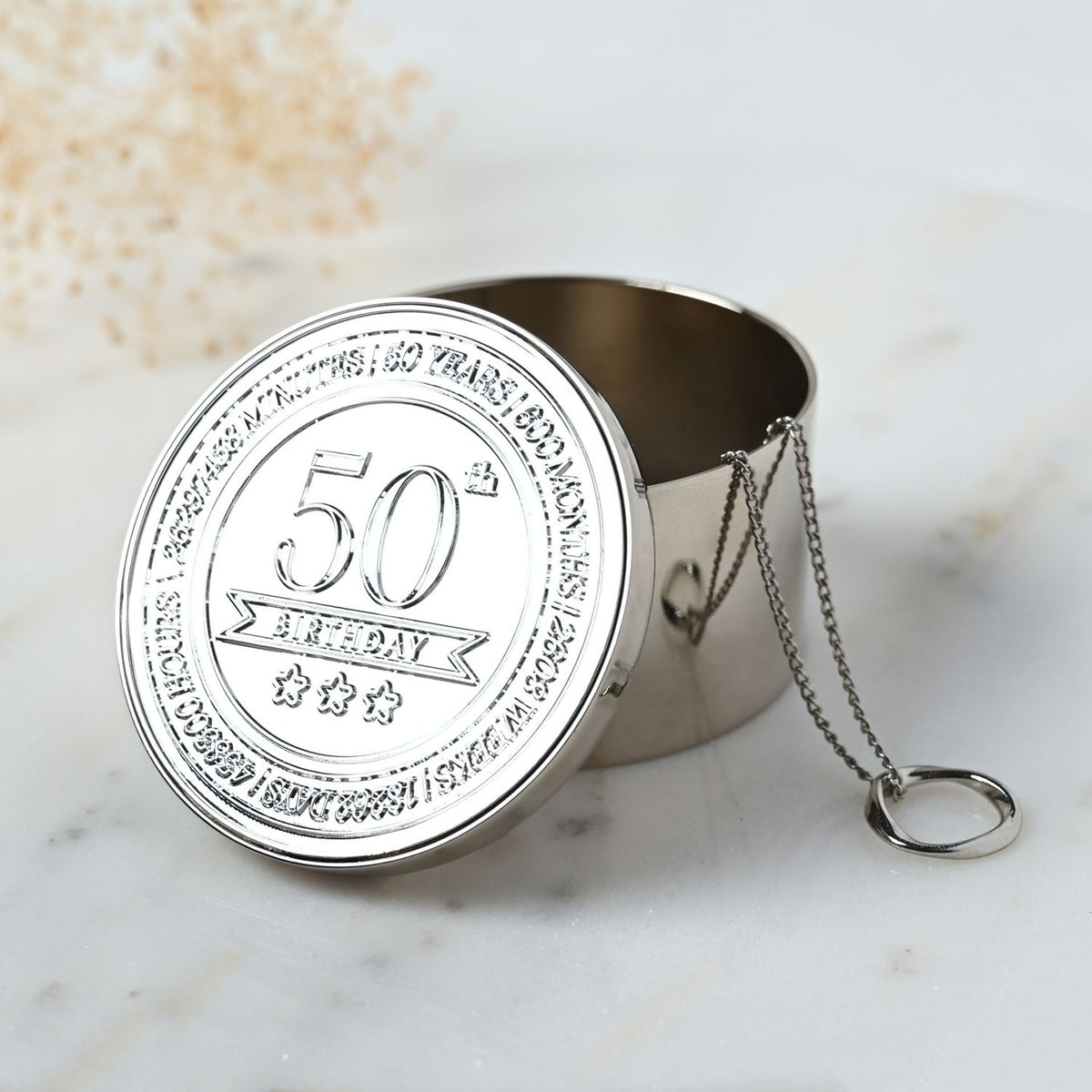 Round silver-toned metal trinket box with an embossed 50th Birthday lid featuring three stars and surrounding text marking years, months, and days, shown open with a chain hinge on marble.