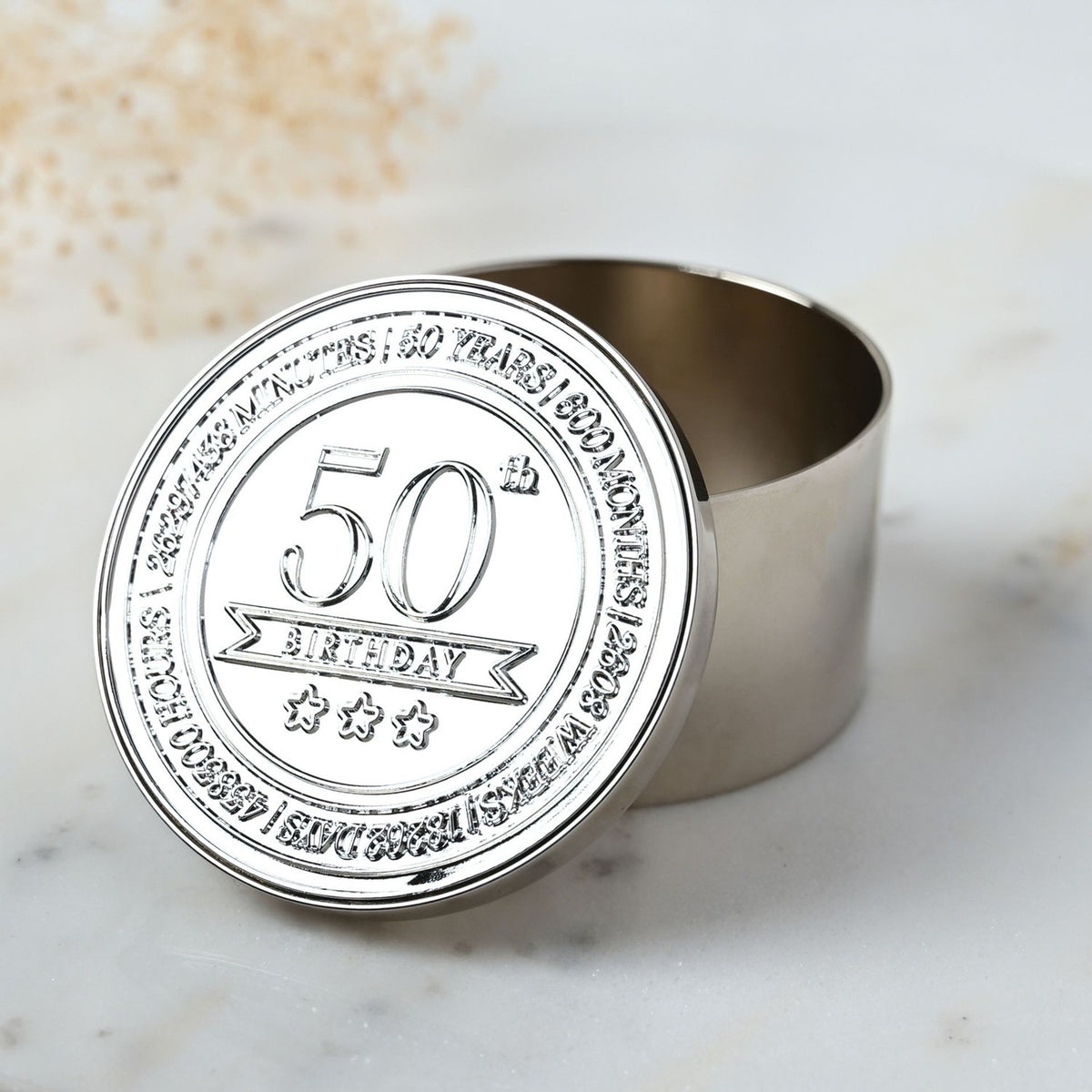 Round silver metal trinket box with embossed lid reading "50th Birthday" and star details, shown open to reveal a polished interior, set on a marble surface.