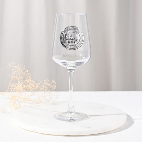 Clear stemmed wine glass with a round silver 40th birthday badge on the bowl, long slim stem and round base, set on a marble tray with a neutral backdrop.